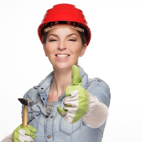 Lady in a red hardhat and green gloves holding her thumb up and holding a hammer