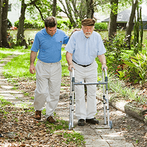 A young man assisting an elderly man with walker walking through garden setting