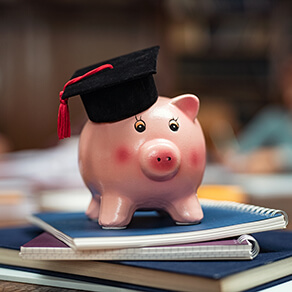 Piggy bank wearing mortar board on pile of books in library while students studying in background. Saving money for college with piggy bank on books in library. Savings and education debt concept.