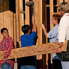 Children and adults carrying a board and working on building a housing frame