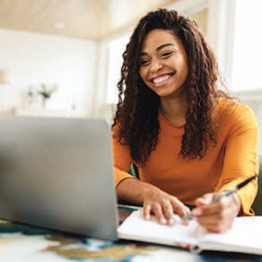 Woman smiling sitting in front of a laptop taking notes