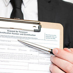 Man holding US tax form W-9 on a clipboard and pointing to it with a pen.